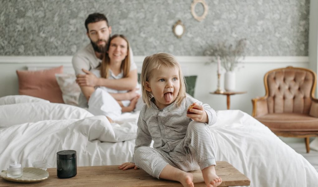 A cute toddler sitting on a bench in a cozy bedroom, with parents sitting on the bed.