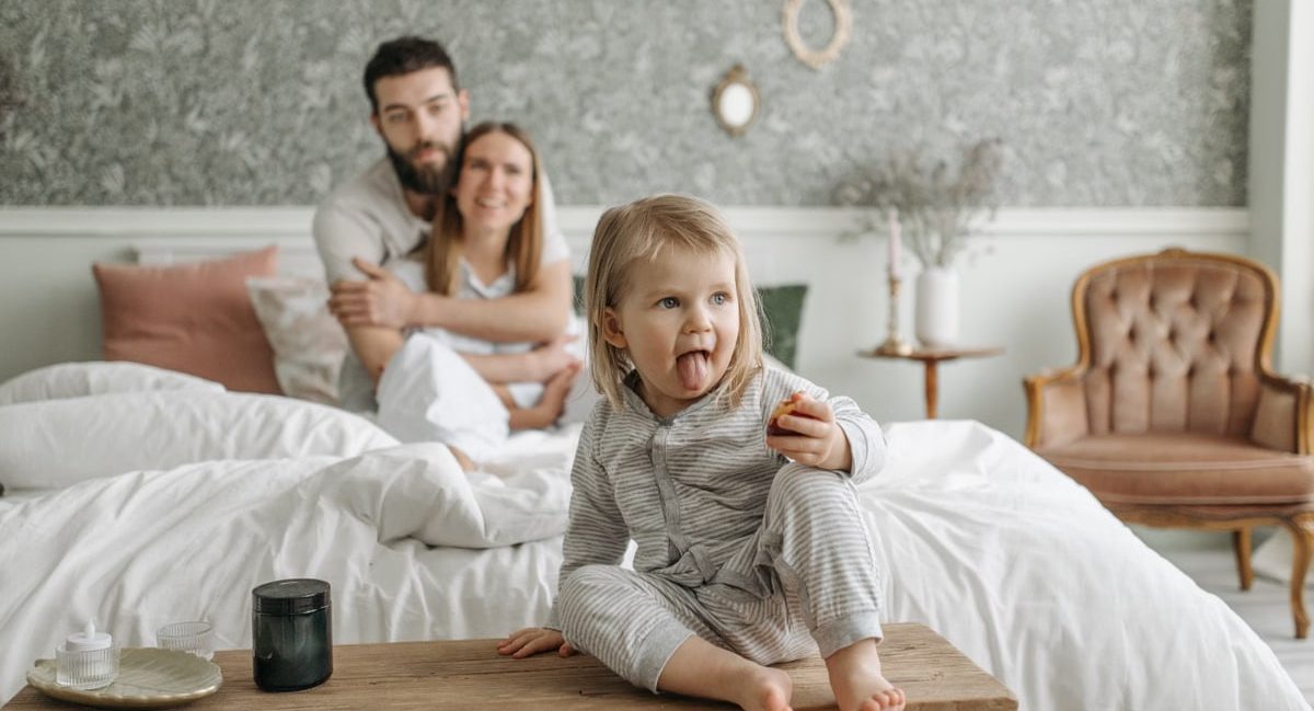 A cute toddler sitting on a bench in a cozy bedroom, with parents sitting on the bed.