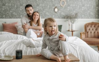 A cute toddler sitting on a bench in a cozy bedroom, with parents sitting on the bed.