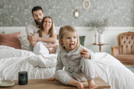 A cute toddler sitting on a bench in a cozy bedroom, with parents sitting on the bed.
